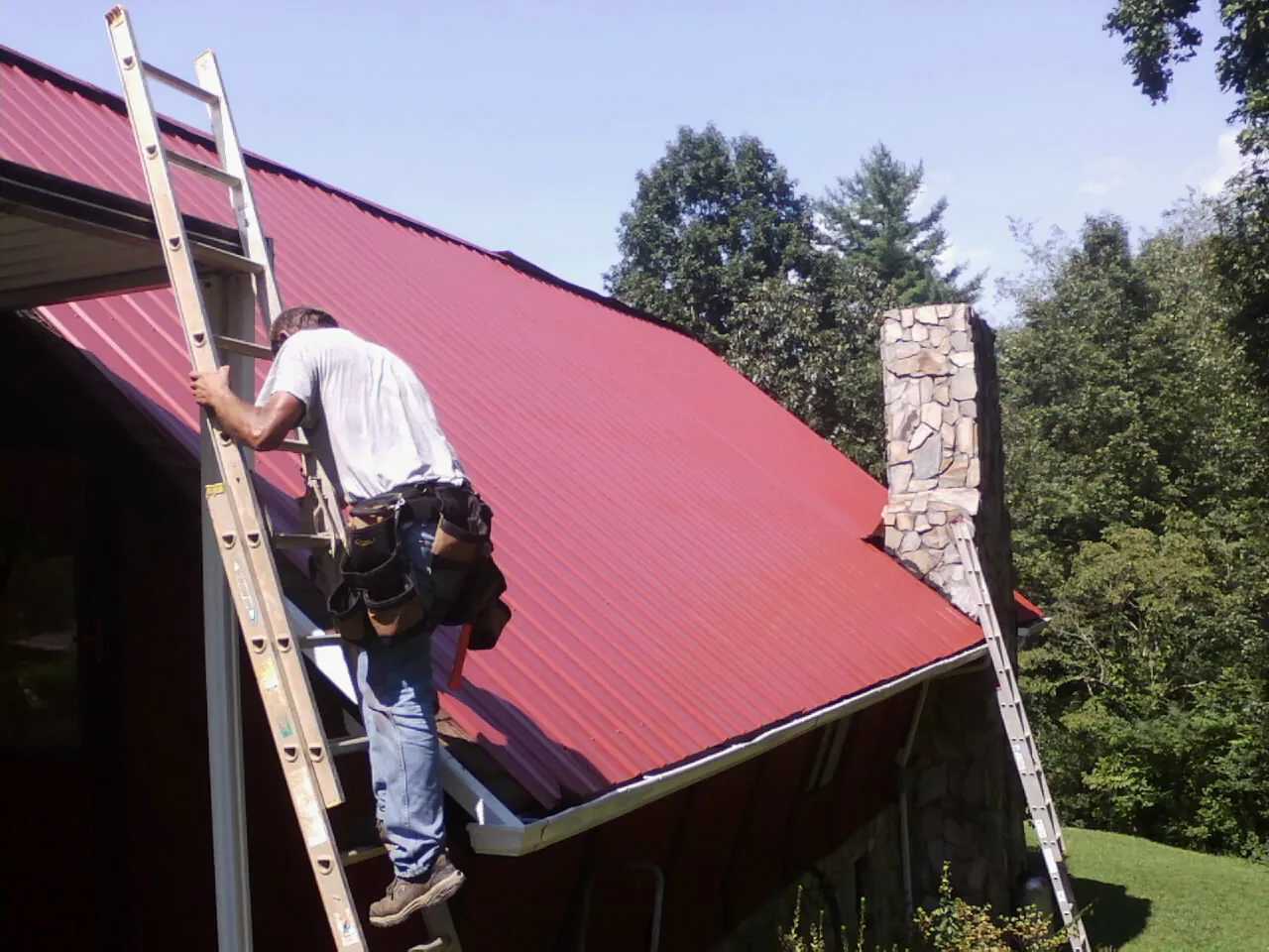 Roofer on a ladder beside a red metal roof and stone chimney — Soffit & Fascia Repair by Roofing Brothers in Olive Branch